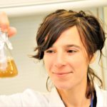 Scientist in a lab coat holds a flask containing an orange liquid, examining it closely in a laboratory setting.