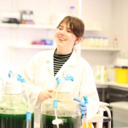 Scientist in white lab coat working with glassware and carboys of green algae in a laboratory setting.