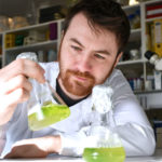 Person in a lab coat holding a conical flask with green liquid in a laboratory setting.